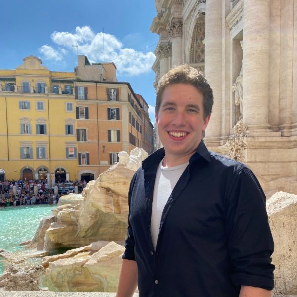 Gabe smiling, in front of a fountain near historic buildings and a crowd of people.