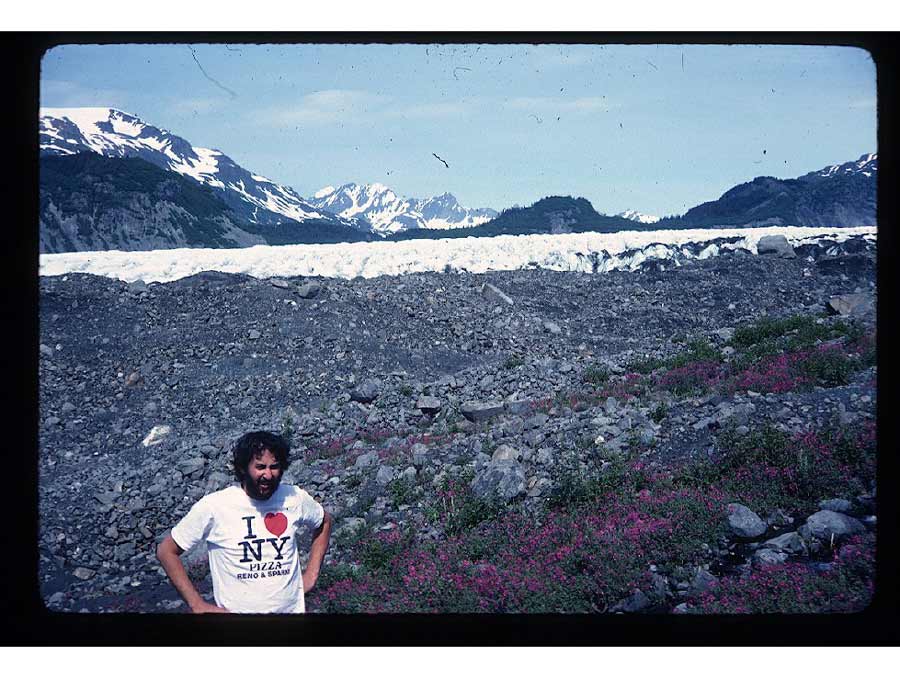 A man in the foreground squinting toward the camera and wearing a "I (heart) NY Pizza Reno & Sparks" t-shirt. Behind him is a gravely hillside abutting a glacier, with large mountains in the distance.