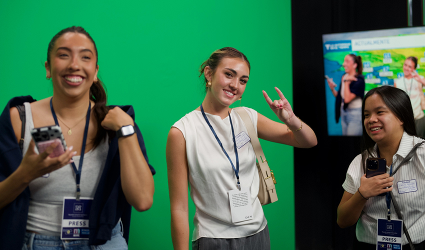 Three students stand in front of a green screen with a weather forecast monitor to the right of them. One holds up the Wolf Pack hand sign.