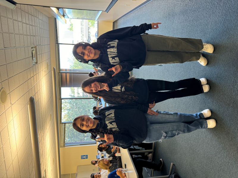 Three people in a classroom setting, each wearing a University of Nevada, Reno hoodie. They are standing and posing with a Wolf Pack hand gesture.