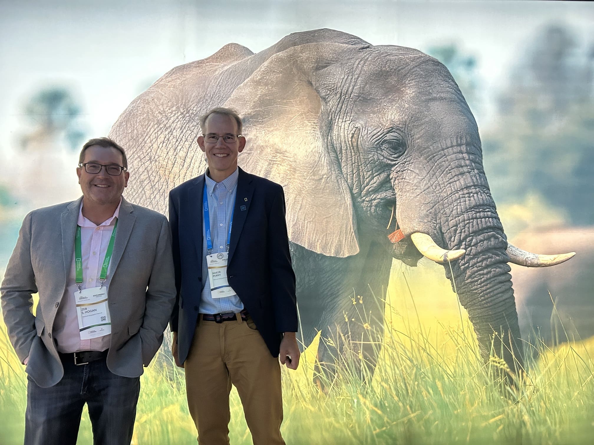 Two faculty members stand in front of photo of an elephant.