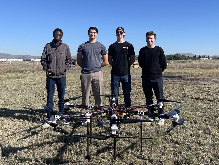 Four young men stand in a field with a drone on the ground in front of them.