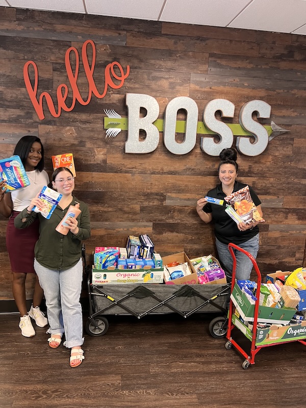 Three people pose while holding food products and hygiene items, around two carts full of more items, in front of a sign that reads "Hello, Boss."
