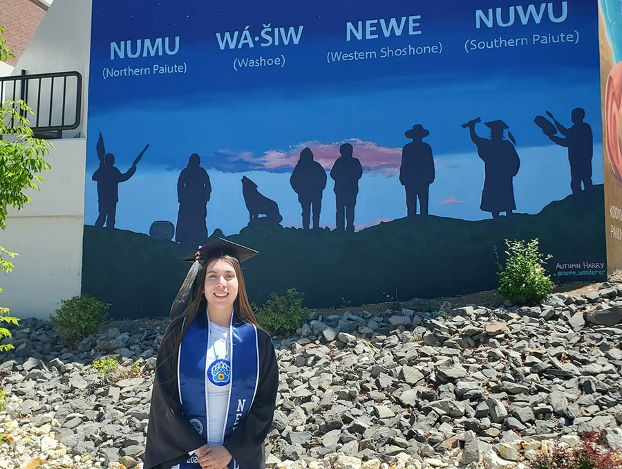A person in graduation attire stands smiling in front of a mural. The mural features silhouettes of Indigenous people under a colorful sky, with labels for the Northern Paiute, Washoe, Western Shoshone, and Southern Paiute peoples.