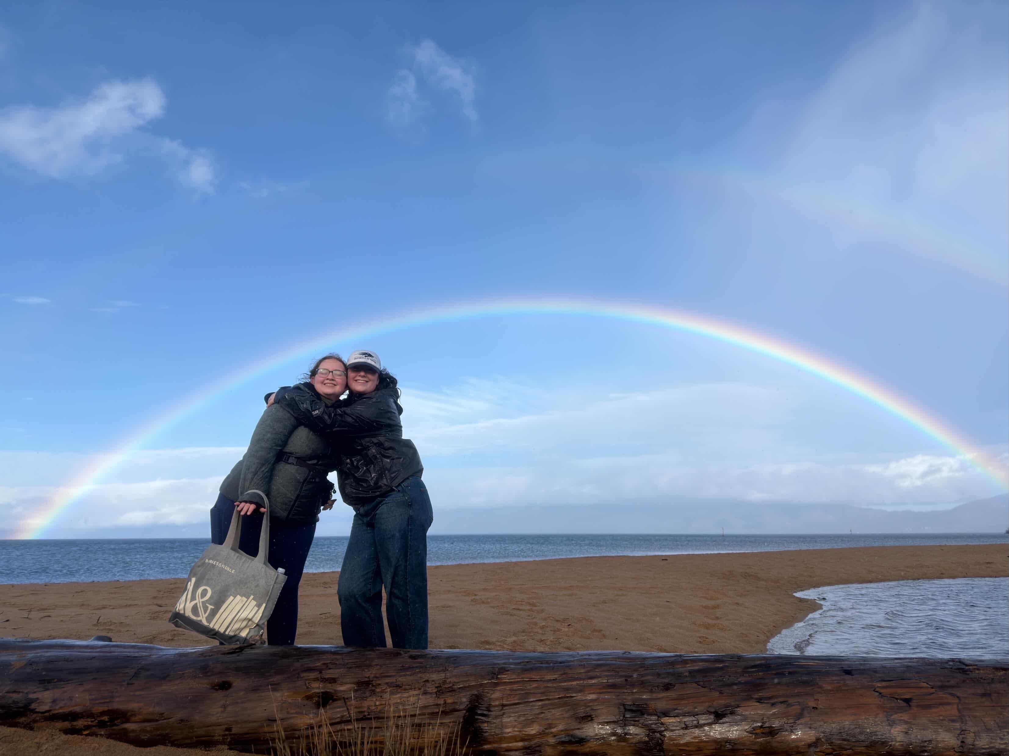 Two students post under a rainbow on the beach at Lake Tahoe.