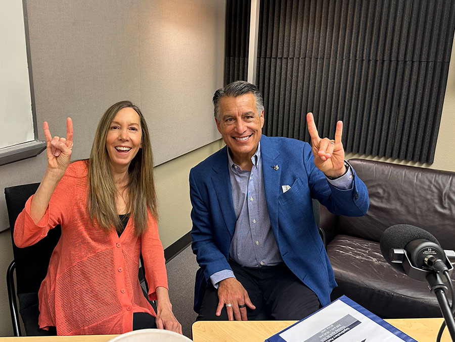 Valerie Fridland sits next to President Sandoval in the podcast recording studio, while making the Wolf Pack hand sign. 