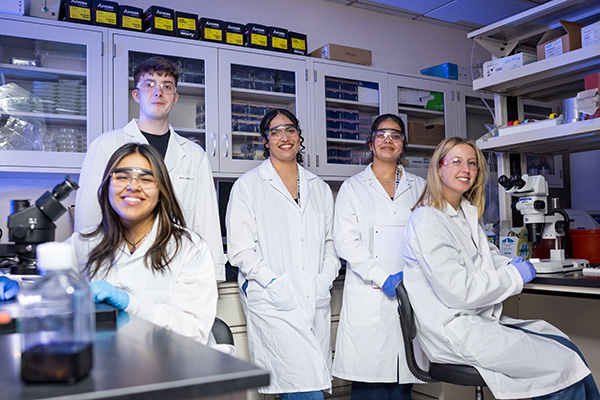 Five students wearing lab coats, safety glasses, and gloves pose together in a research laboratory surrounded by microscopes, scientific equipment, and laboratory supplies.