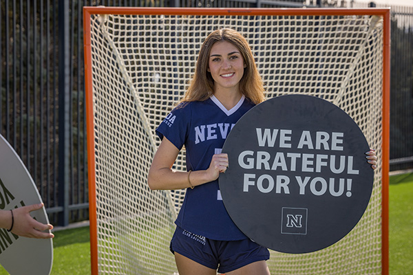 A student-athlete from the Nevada women&rsquo;s lacrosse team stands in front of a lacrosse goal holding a sign that reads &ldquo;We Are Grateful for You!&rdquo; during the University&rsquo;s Giving Day campaign.