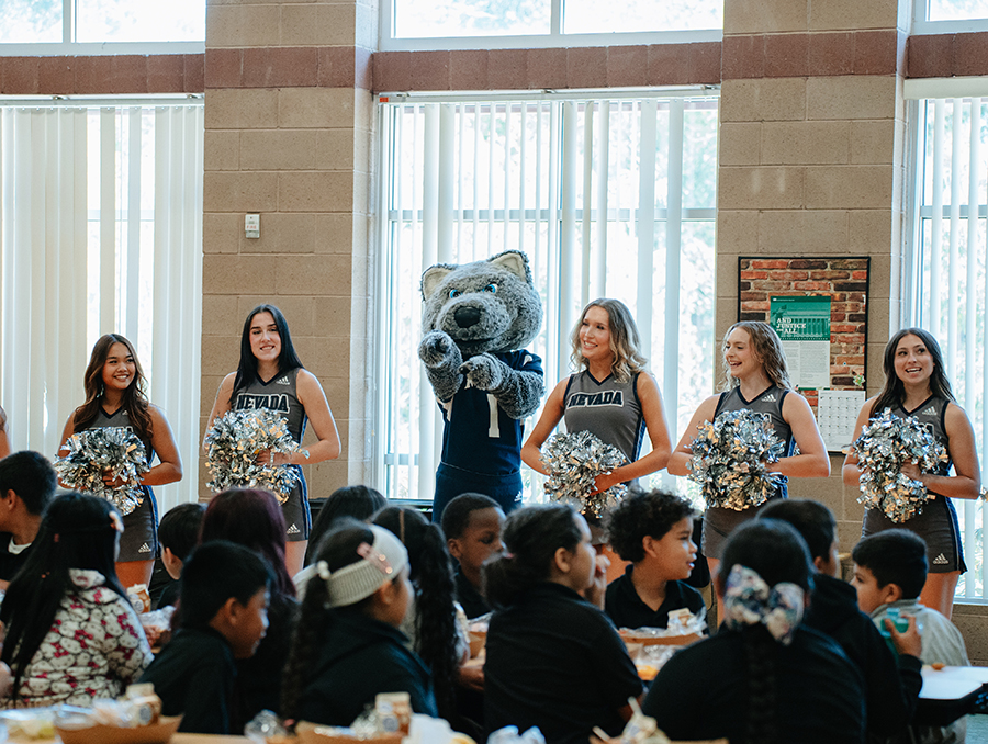 Mascot Alphie Jr. pointing at seated schoolchildren with Nevada cheerleaders.