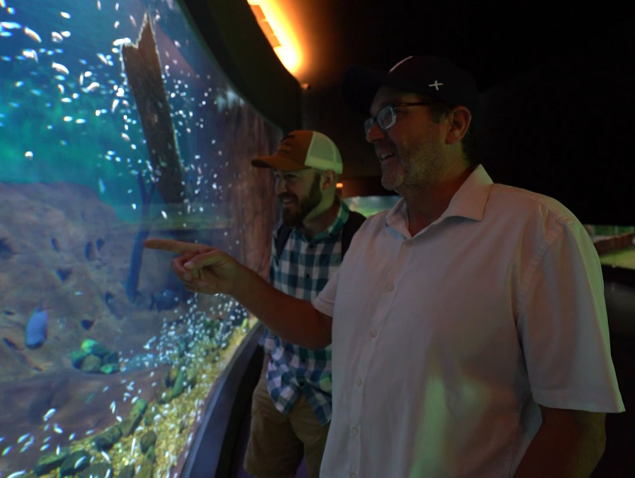 Two men standing in front of the glass in an aquarium, both smiling and one pointing at something in the tank.