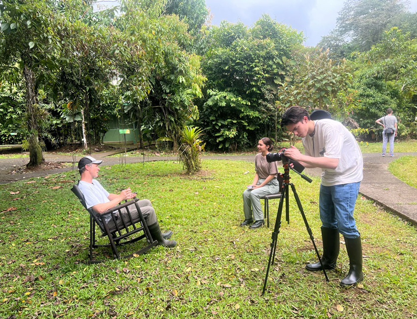 A student using a camera to record an interview between two people in a lush, green environment in Costa Rica.