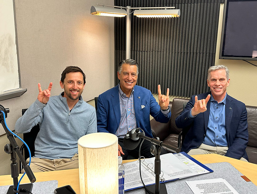 Russ Renzas sits next to President Sandoval who sits next to Dean Tom Weller in the podcast recording studio, while making the Wolf Pack hand sign. 