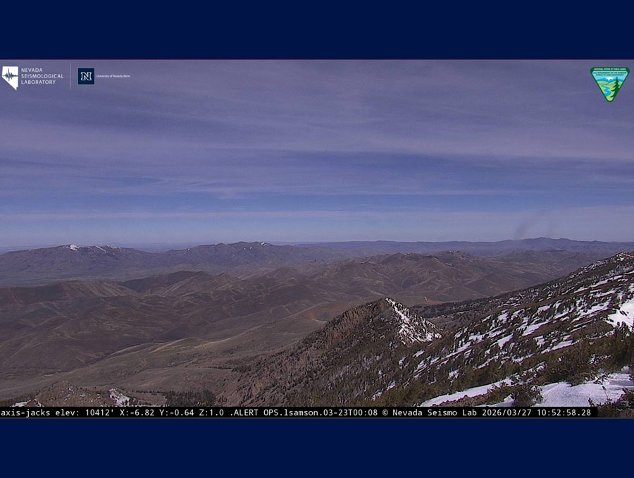 A camera feed with logos at the top including the Nevada Seismological Laboratory, the University of Nevada, Reno and the Bureau of Land Management. Information text is in a banner across the bottom of the feed. The feed shows several mountain ranges, most of which have little snow on them.