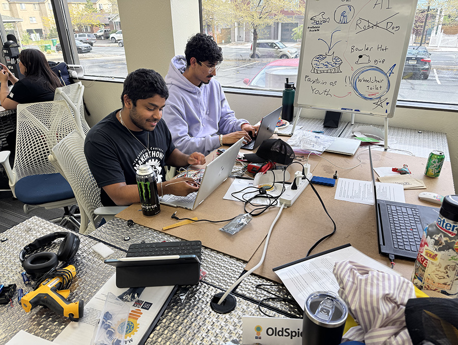 A small group of people working at a cluttered table with laptops, drinks, and electronic equipment with a large whiteboard in the background with drawings and notes.