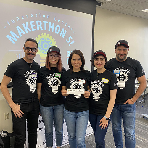A group of five people standing in front of a drop-down presentation screen with the words, "Makerthon 5!" A person holds a sign that says "The Dream Team."