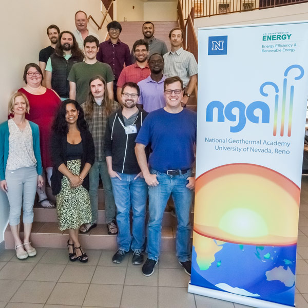 A group of 14 people smile for a photo on a flight of stairs next to a banner with a graphic of the Earth and its interior and text that reads, "National Geothermal Academy, University of Nevada, Reno"