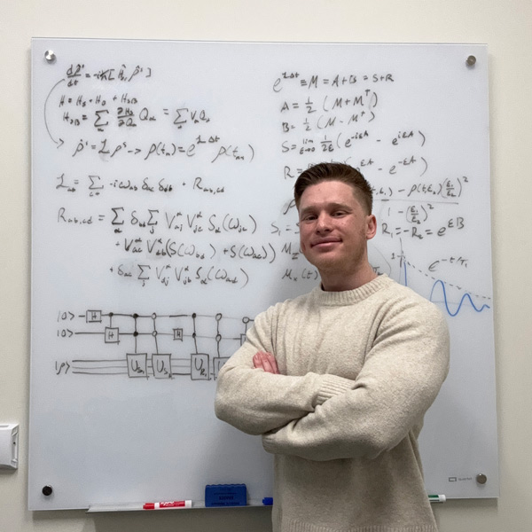 Noah Huerta smiles in front of a whiteboard with equations and diagrams.