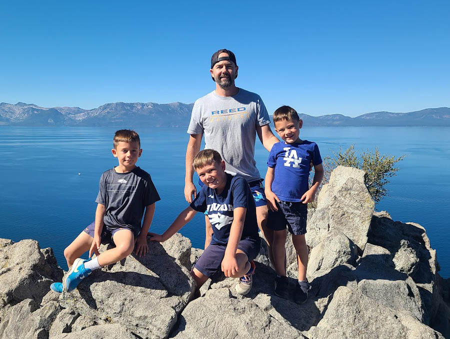 A man and three young boys in casual wear stand on rocks with a lake in the background.