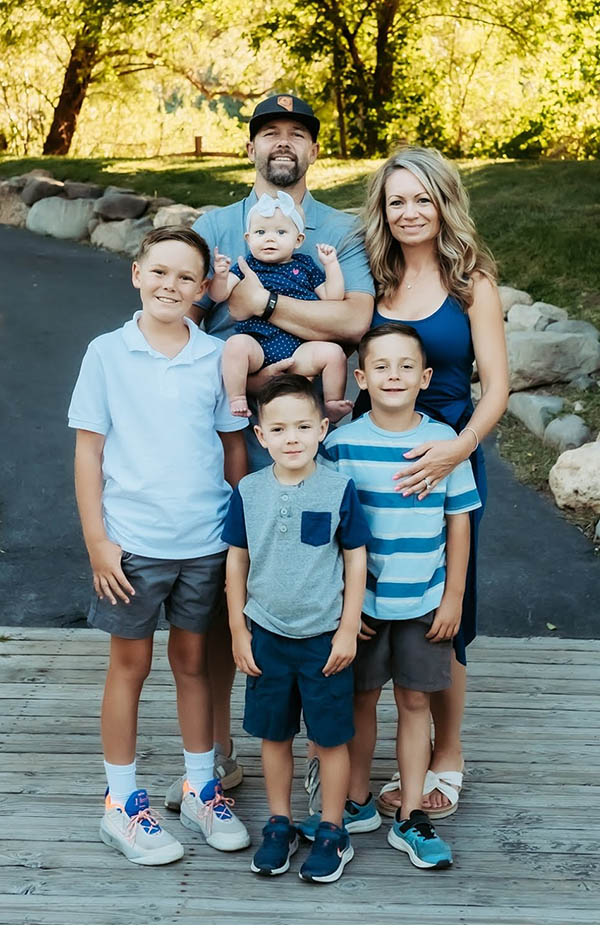 A man holding a baby stands outdoors next to a woman; three boys stand in front of them. 