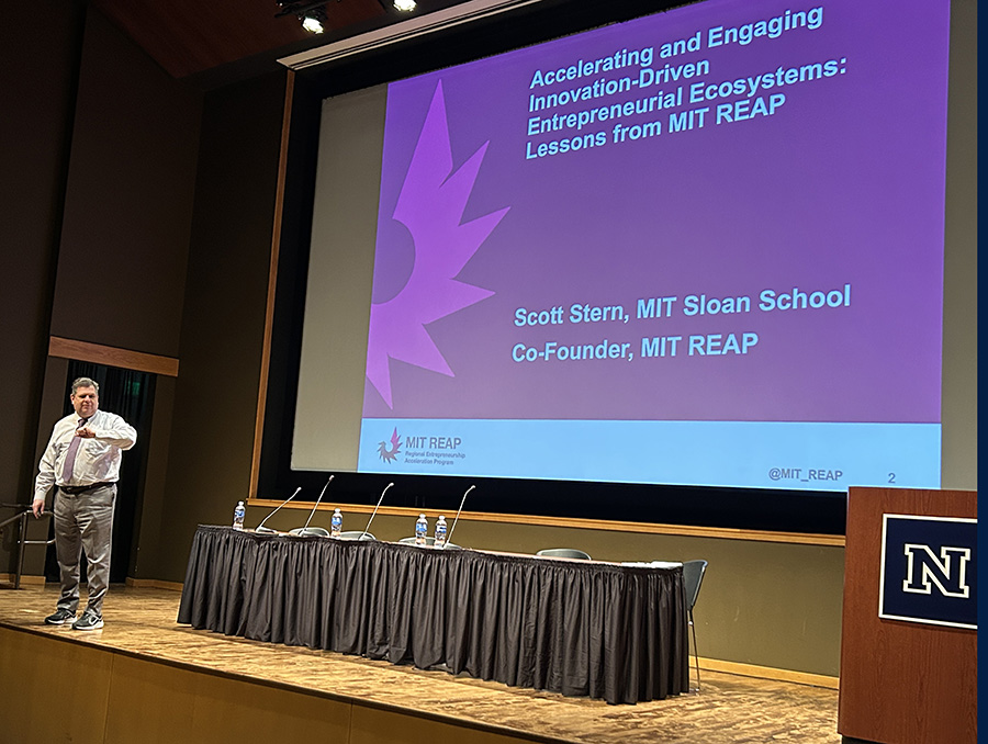 Scott Stern from MIT Sloan School on a stage next to a presentation slide on stage giving a presentation for Research to Reality symposium..