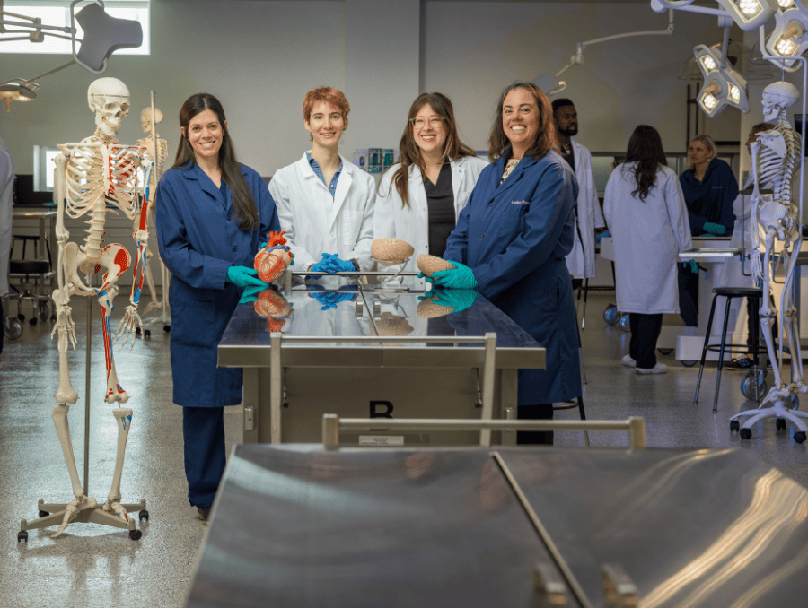 Two instructors and two students standing next to an anatomy table in a lab.