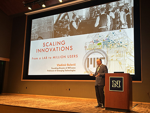 Dr. Vladimir Bulović of the MIT School of Engineering stands on a stage in front of a large screen giving a presentation.