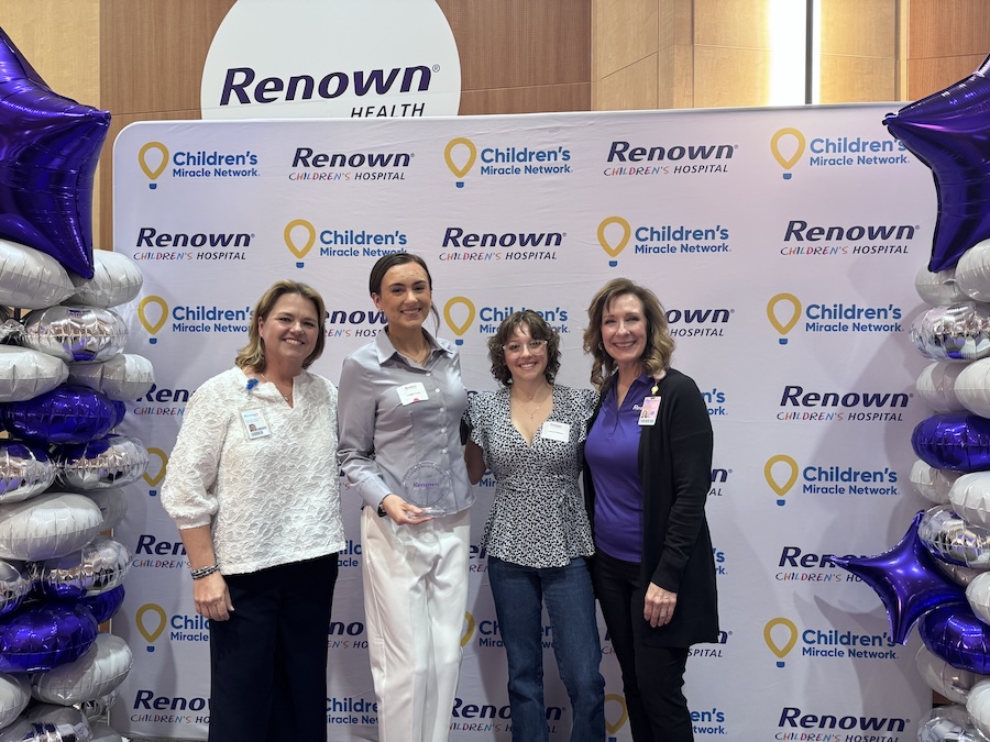 A group of four people pose together in front of a Renown Hospital step and repeat banner.