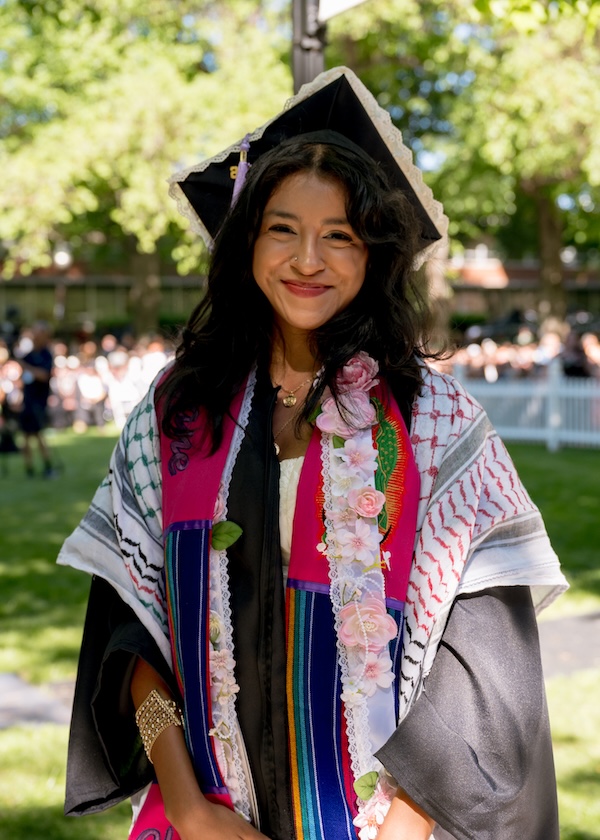 A smiling graduate in cap and gown with a flower sash smiles at the camera.