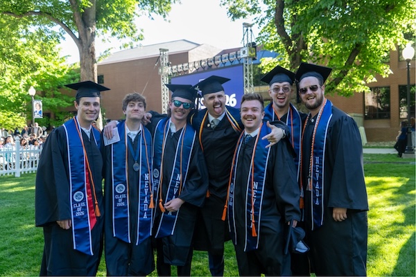 A group of joyful graduates smiles near the stage in cap and gowns.