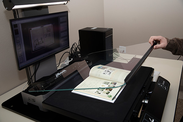 Overhead book scanner in a digitization lab capturing an open magazine, with a computer monitor displaying the scan preview and a hand lifting the scanner lid.