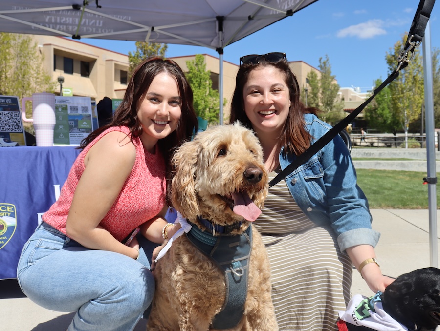 Emma poses with Madison in street clothes, outside, while petting a golden doodle dog.