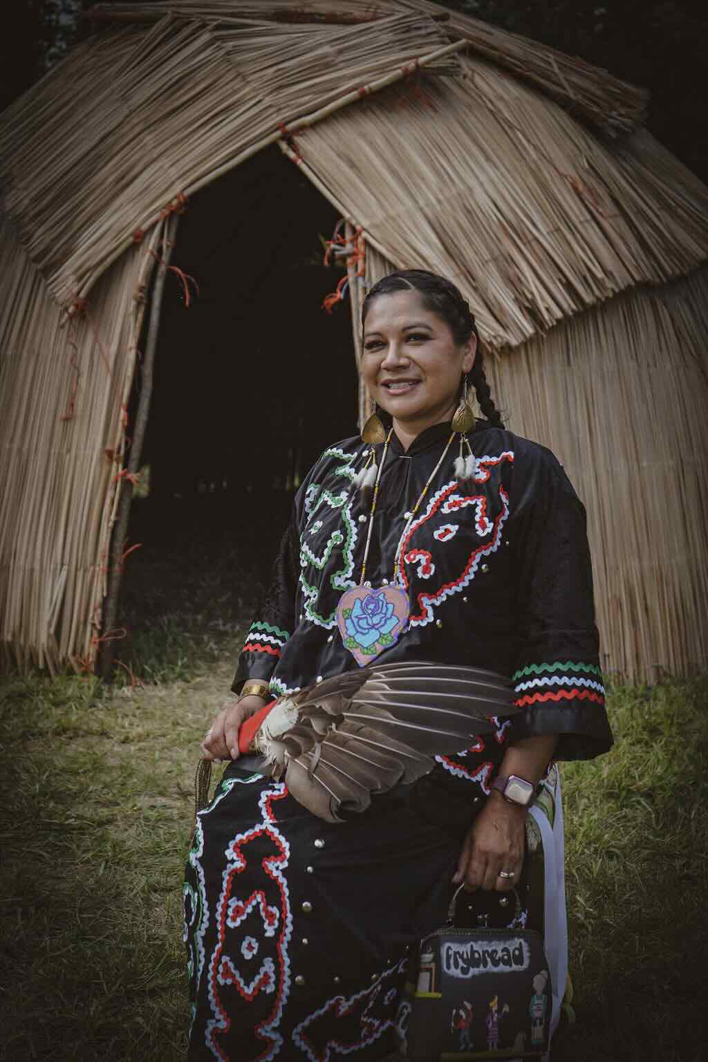 Misty Youngbear in traditional dress sitting outside of a straw hut.