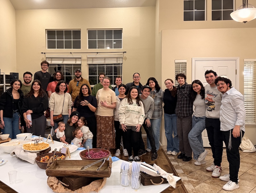 A large group gather in a dining room, smiling in front of a table with food.
