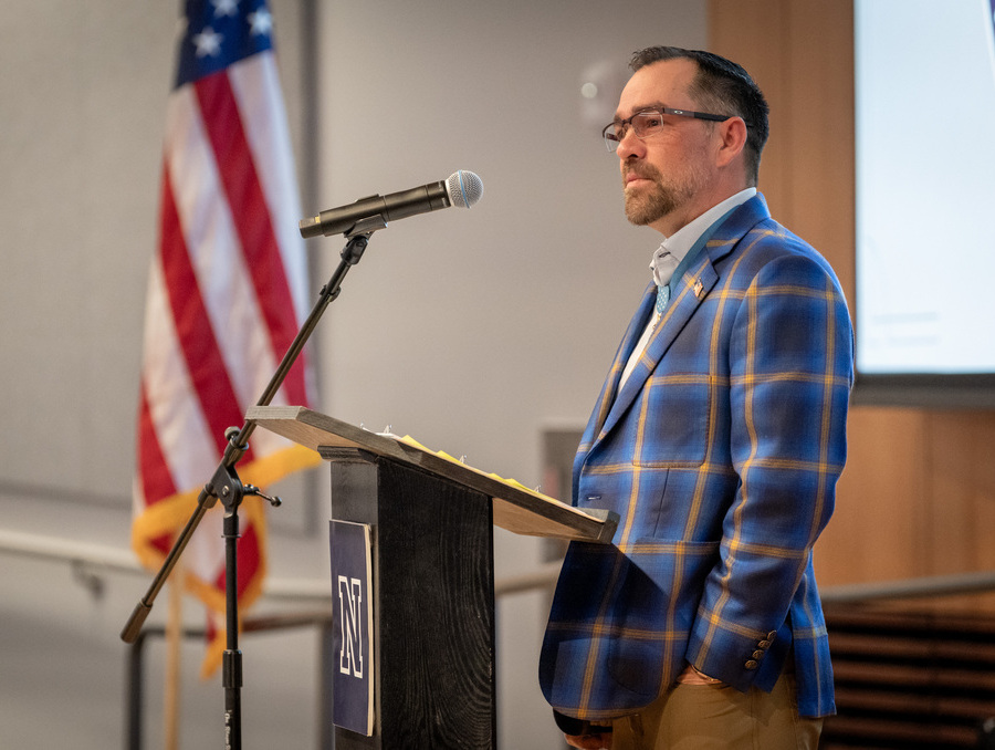 Clint Romesha stands at a podium speaking during the ENGAGE event, with a microphone and U.S. flag visible behind him.