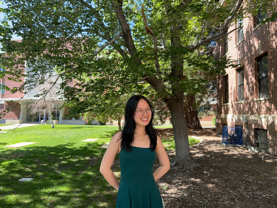 Young woman in green dress poses outdoors with a tree buildings behind her.