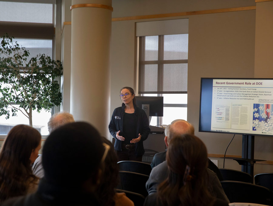 Jennifer Wilcox speaks to an audience in the JCSU Great Room with a screen next to her with text reading, "Recent Government Role at DOE."
