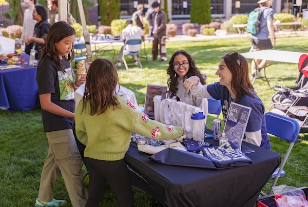 Two people smiling while working a table at an event with two other people talking and learning from them. 