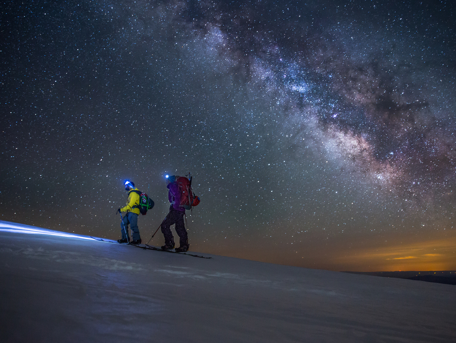 Two mountaineers at night on the side of a snowy mountain, walking in the dark with headlamps. The night sky, full of radiant starts, is illuminated behind them.