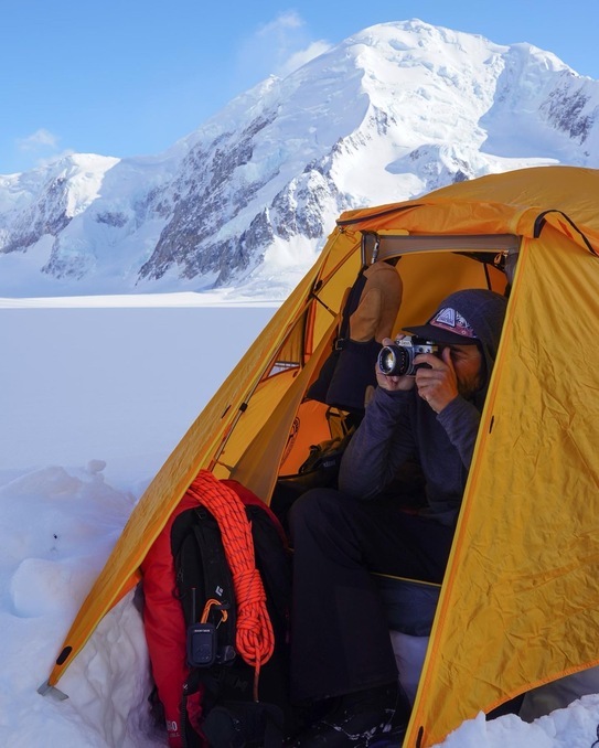 Cahill in a tent on the side of a snowcapped mountain with his camera in hand, capturing a photo.