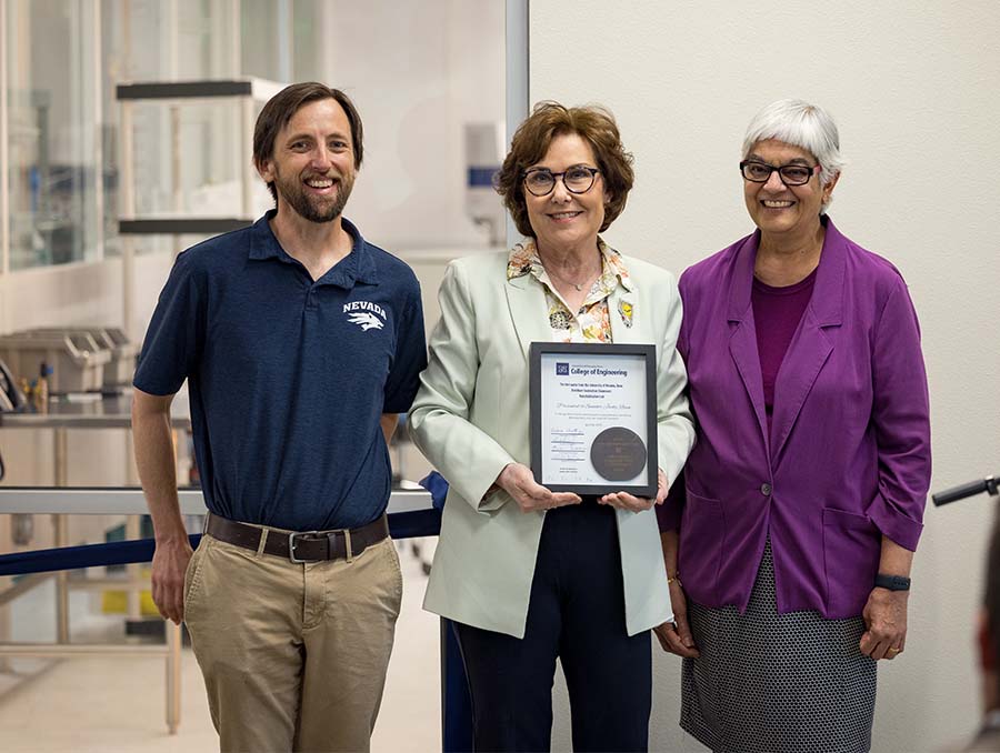 Three people stand indoors with a window to a lab in the background; the middle person is holding a framed certificate.