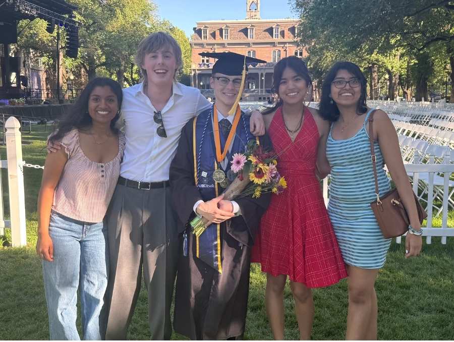 Five people, including Riley Gillis in the middle wearing graduation regalia, smile on the University Quad.