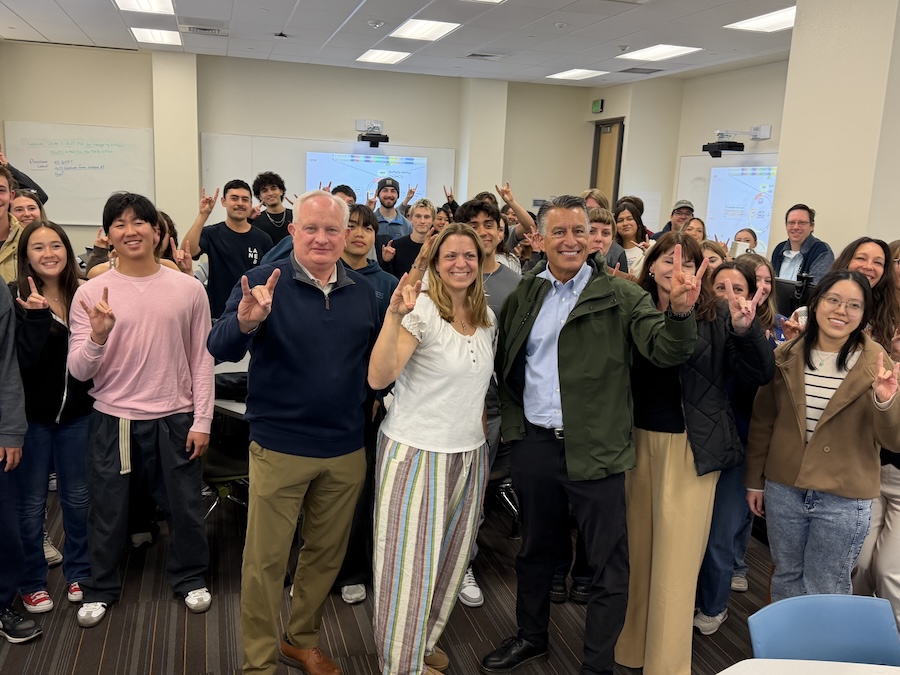 Pamela stands between Provost Thompson and President Sandoval inside her classroom, smiling and receiving the award. They all hold up wolf pack hand signs, and the class is full of her smiling students, posing behind them.