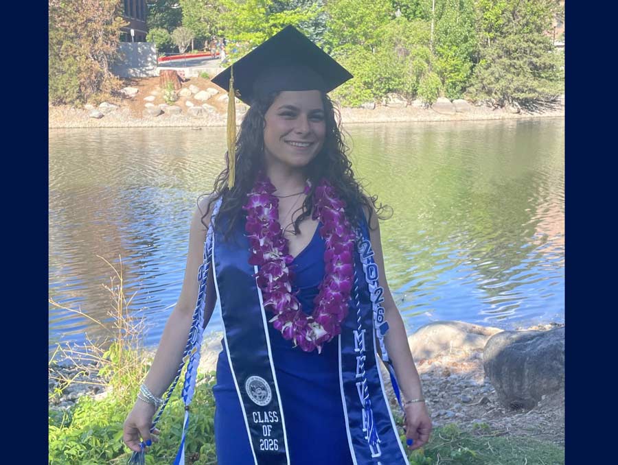 A young woman wearing a graduation cap and stole, lei and cords, smiling for a photo in front of Manzanita Lake.