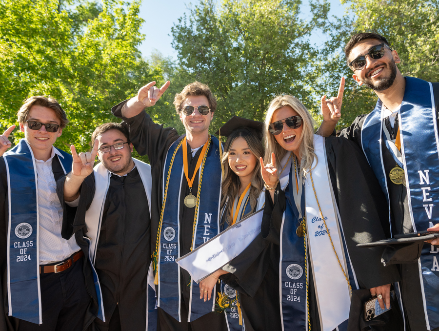 Students pose in graduation attire on the quad, holding up wolf pack hand signs and smiling.
