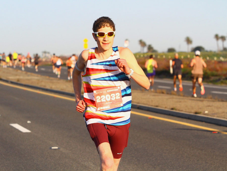 A man, wearing running shorts and sunglasses, running on the road with other runners in the background. 