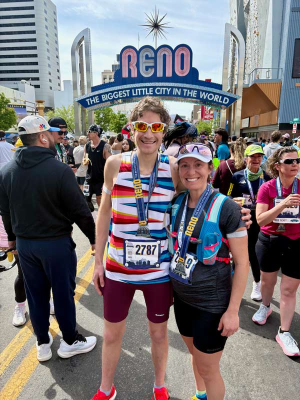 Daniel Trugman hugs a woman, both wearing medals, race bibs and running gear, in a crowd of people under the Reno Arch.