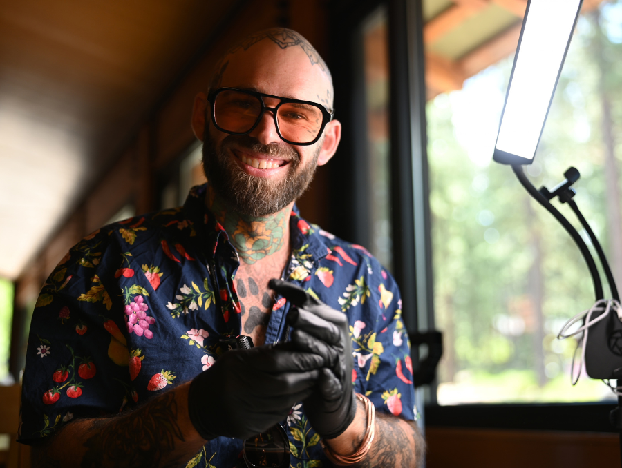 Nick Giordano smiles while wearing orange-tinted glasses, a patterned shirt and black gloves, holding a tattoo machine beside a window.