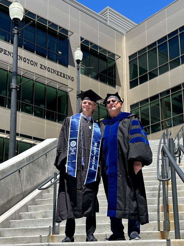 Two people smiling while wearing graduation attire on the steps outside a building.
