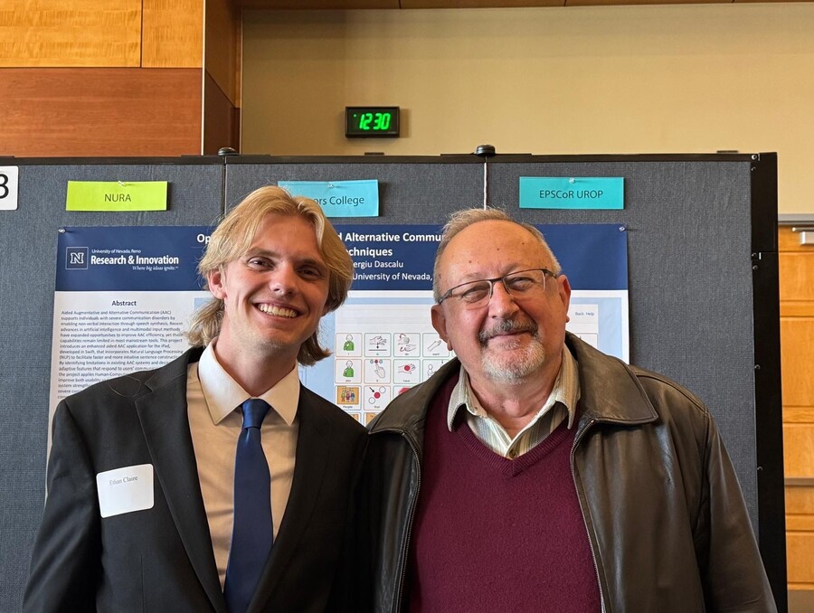 A smiling young man in a dark suit stands beside an older man in glasses and a leather jacket, posing before a university research poster.