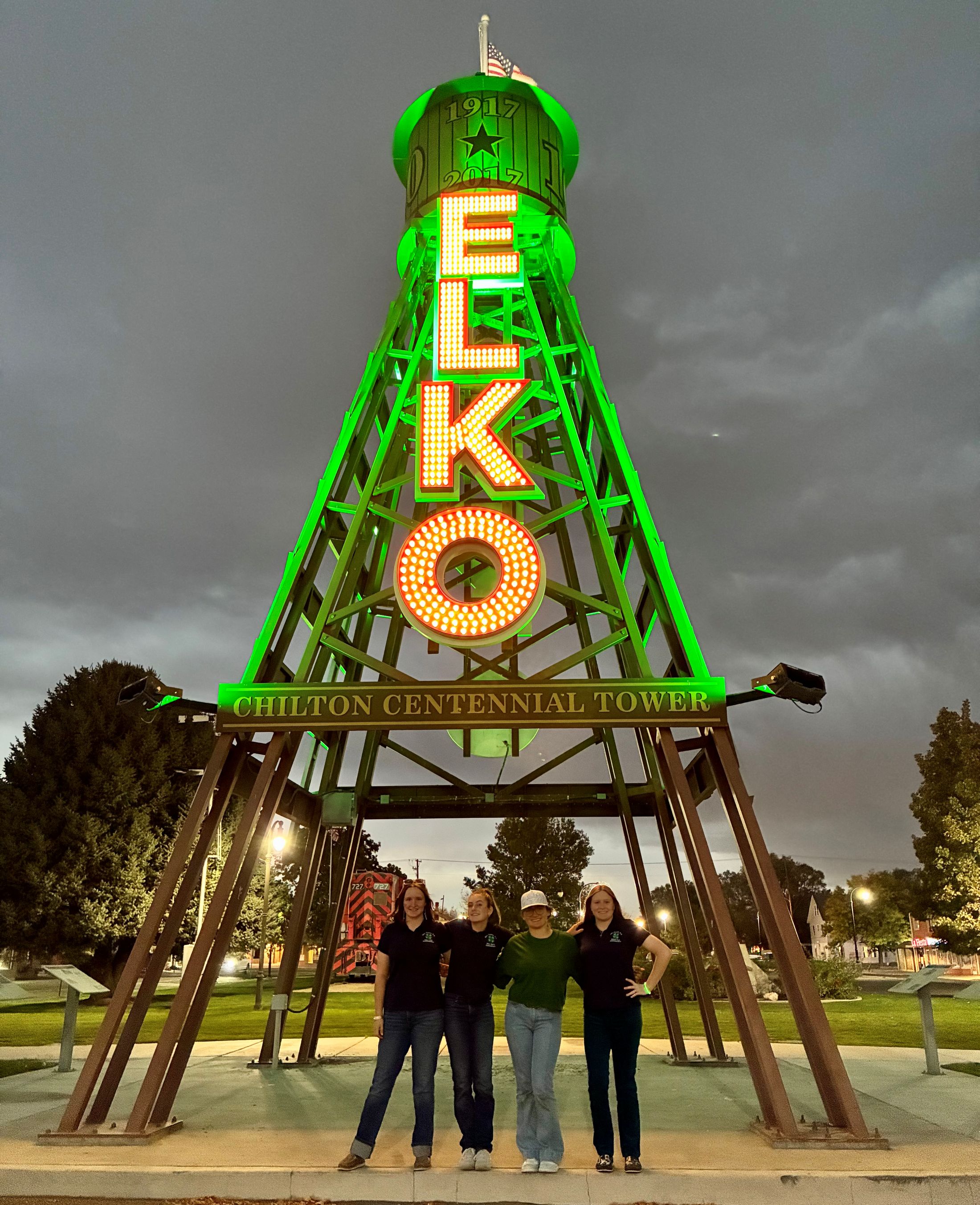 A lit neon sign of Elko's Chilton Centennial Tower with four people standing under it, smiling.